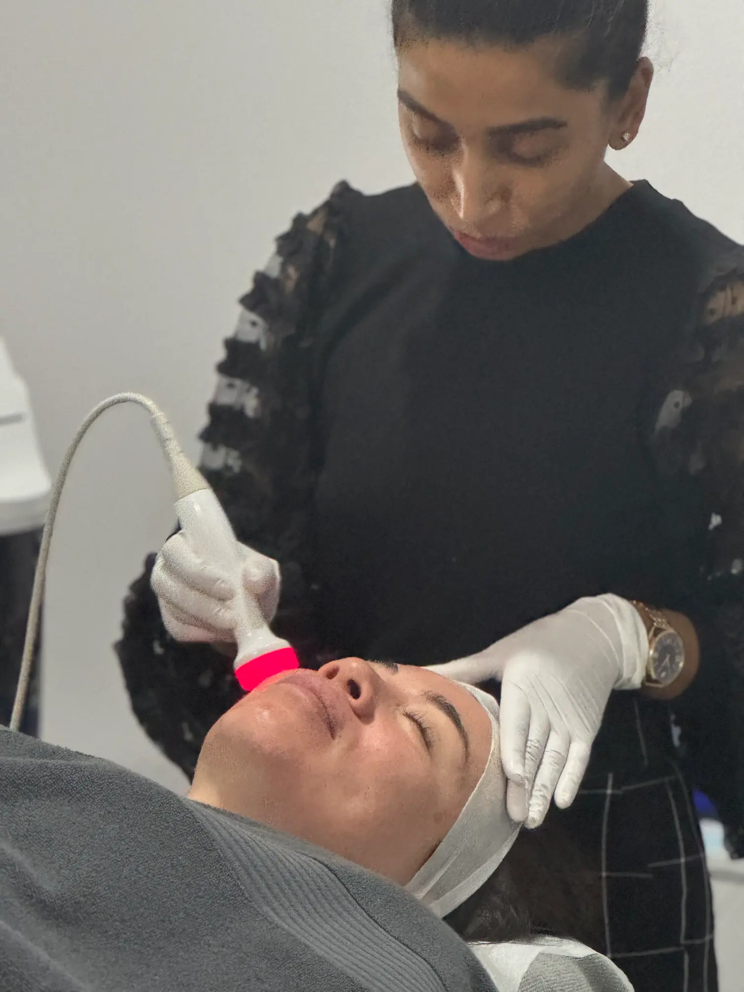 Anushka is shown performing a skin resurfacing treatment on a client. The client is lying down with a headband on, and Anushka, wearing gloves, is using a device that emits red light to improve the texture and appearance of the client's skin through skin resurfacing
